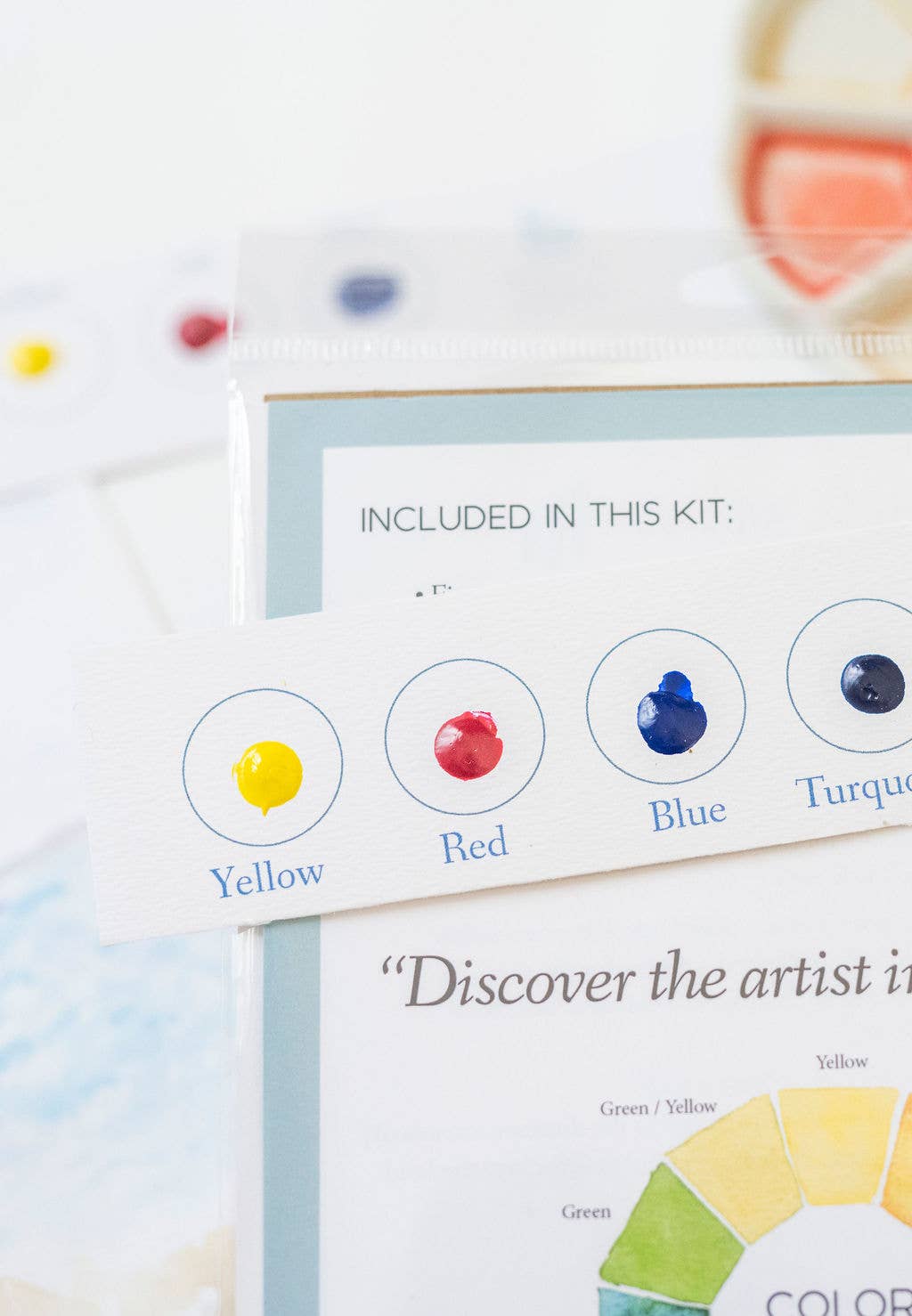 A close-up of a color discovery art kit showing printed color cards with labeled dots for Yellow, Red, Blue, and Turquoise, plus a partial color wheel on a white booklet.