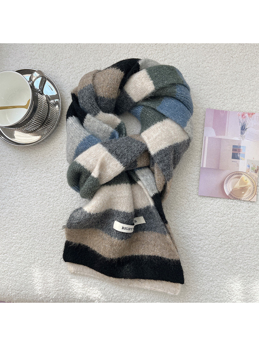 A chunky knit scarf with a large checkered pattern in beige, gray, blue, and black, styled on a textured white surface next to a silver cup and a small card.