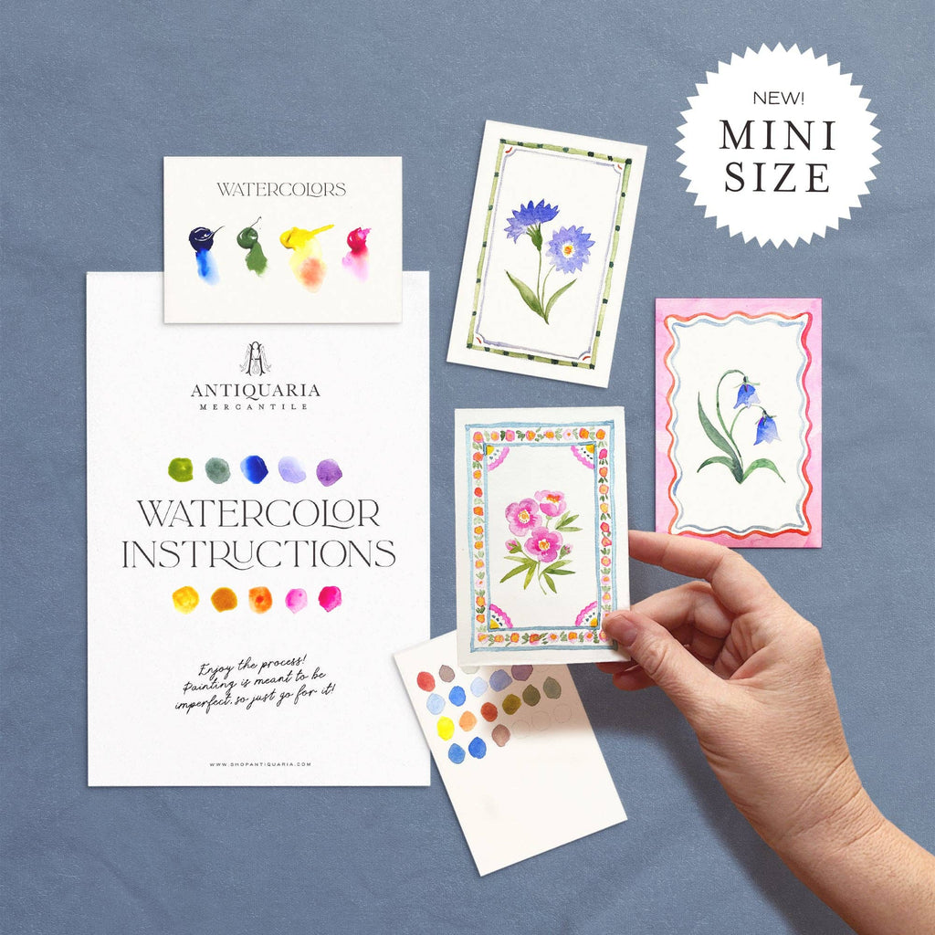 Hand holding a mini watercolor card with floral design on a blue background, featuring Antiquaria Merchanteria branding.