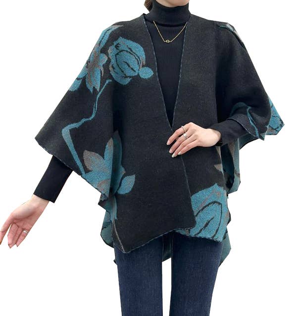 A woman wearing a black open-front poncho-style cardigan with a teal floral pattern, layered over a black turtleneck and blue jeans against a plain white background.