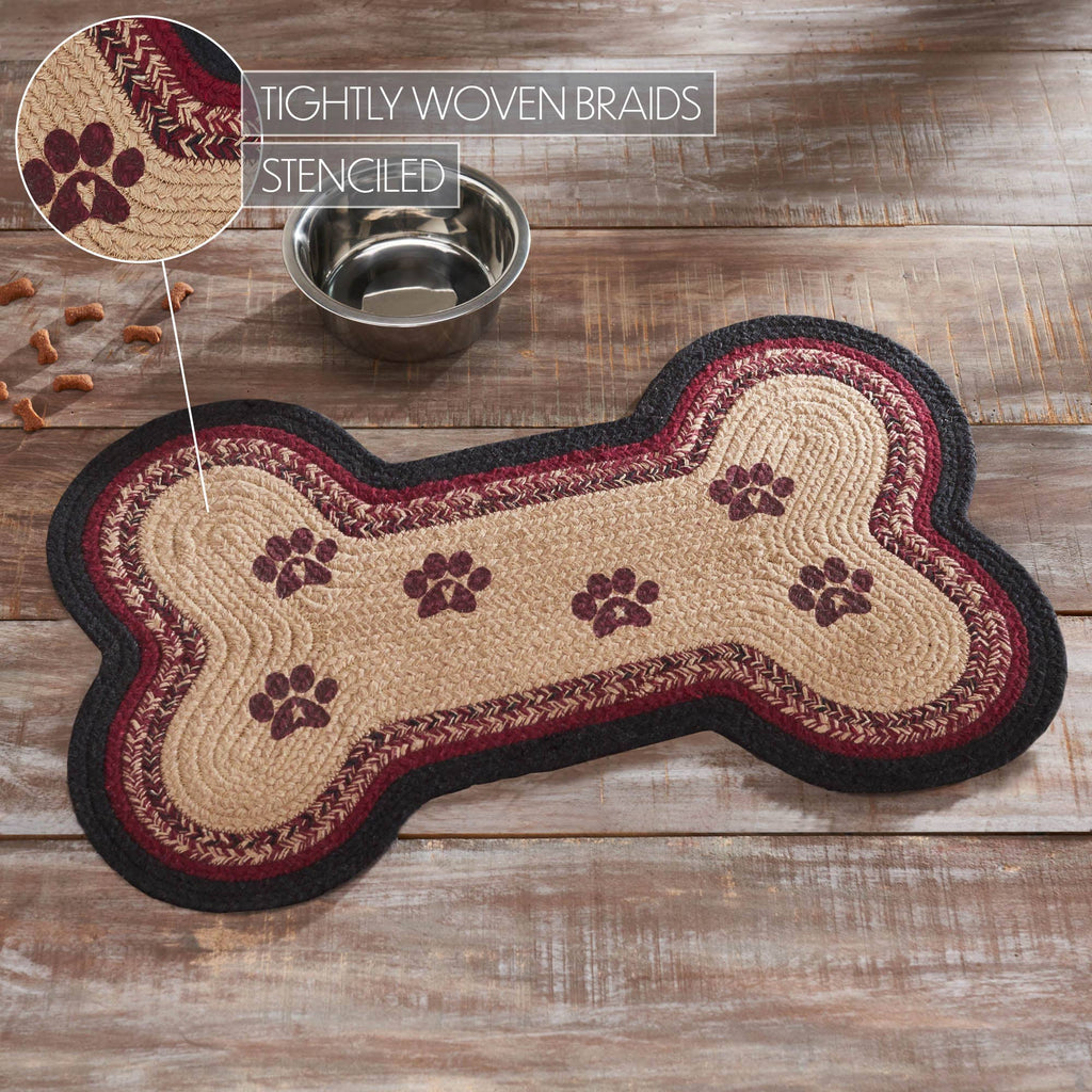 A bone-shaped braided rug with a tan center, burgundy and black braided borders, and dark brown paw print motifs, shown on a rustic wood floor beside a metal bowl. An inset callout highlights the tightly woven braids and stenciled paw prints.
