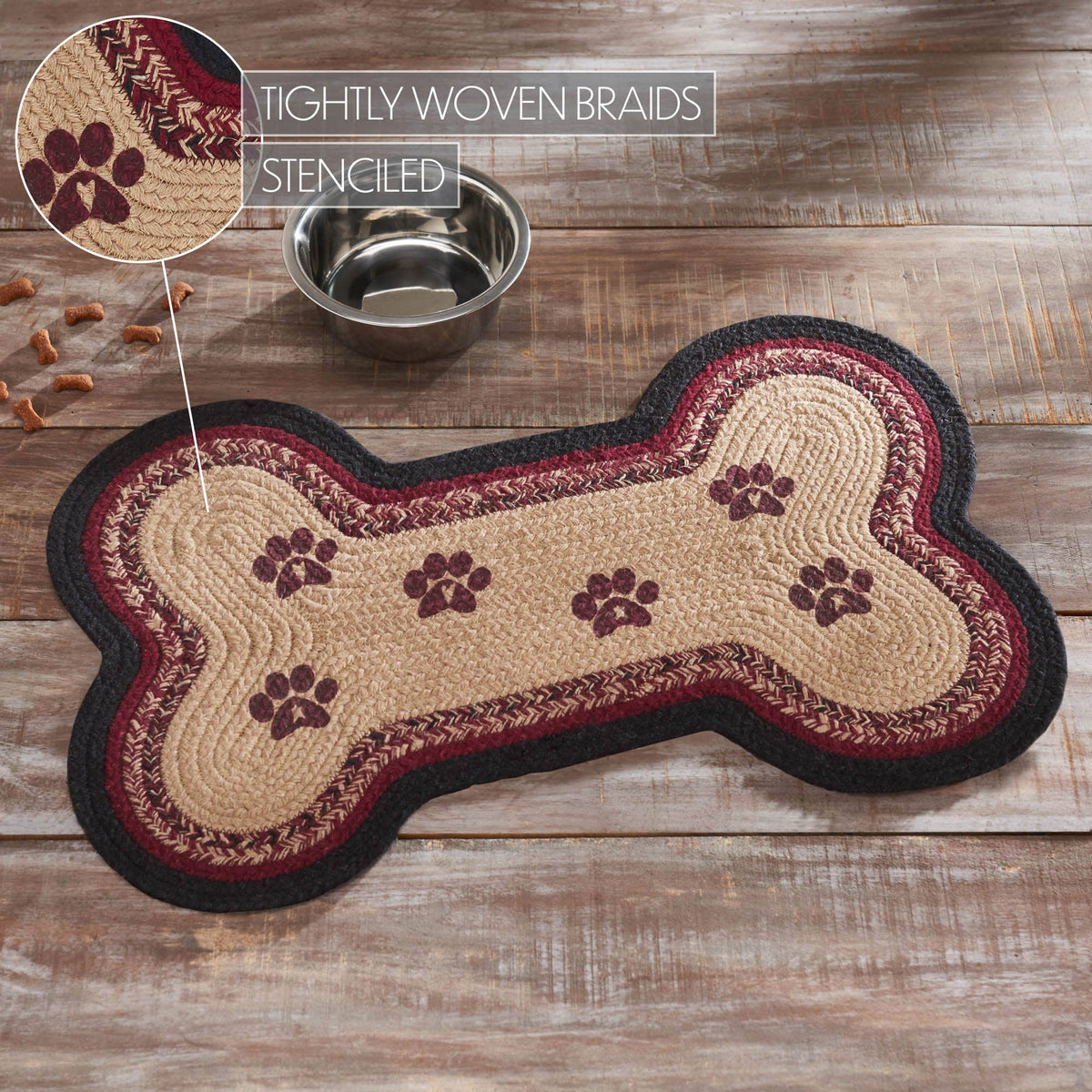 A bone-shaped braided rug with a tan center, burgundy and black braided borders, and dark brown paw print motifs, shown on a rustic wood floor beside a metal bowl. An inset callout highlights the tightly woven braids and stenciled paw prints.