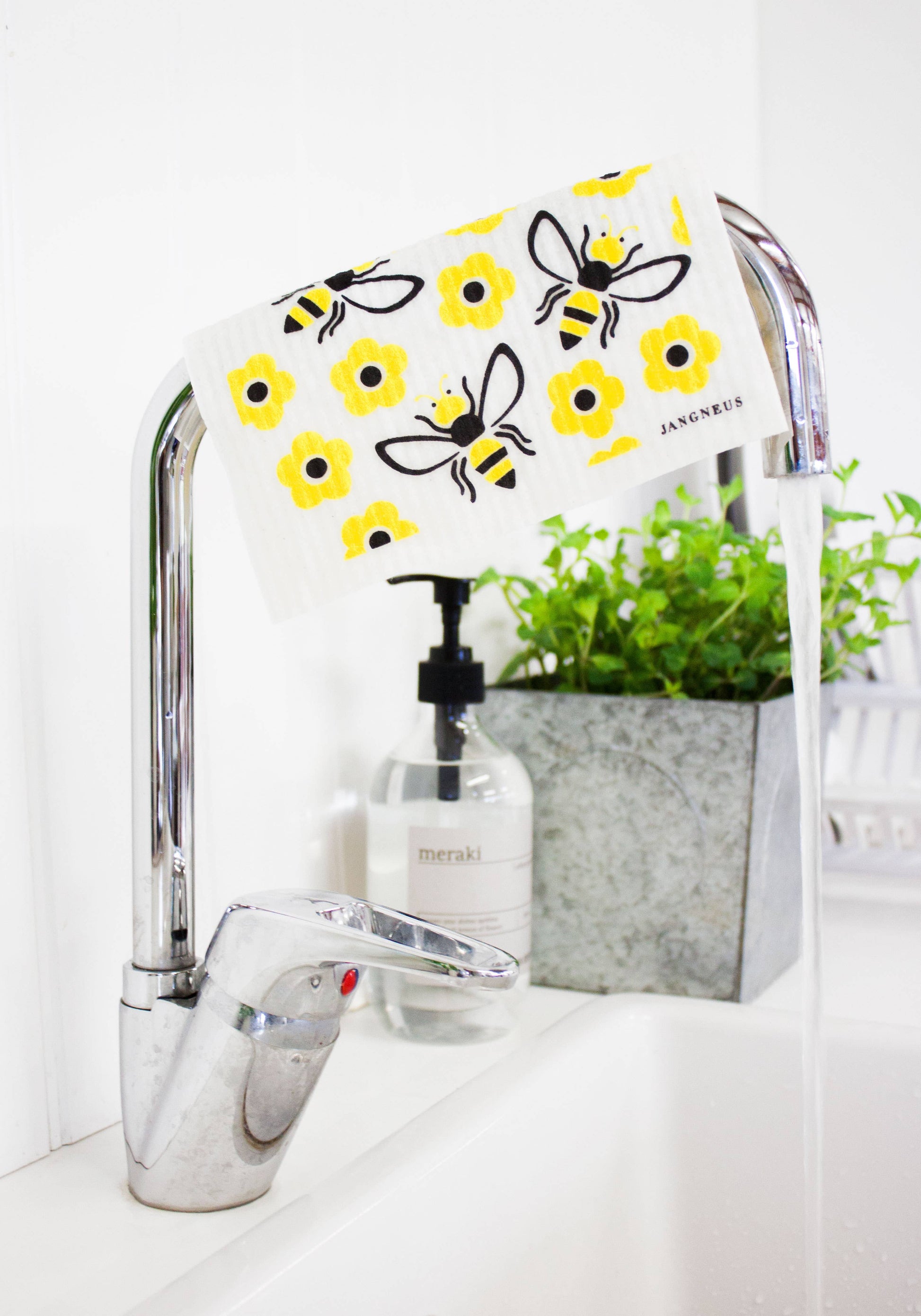 Chrome faucet with a decorative cover featuring bees and flowers, next to a bottle of soap and a plant on a white surface.