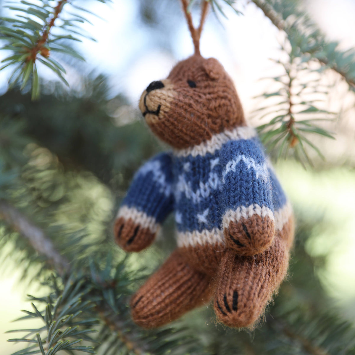Knitted teddy bear ornament wearing a blue sweater with white patterns, hanging on a tree branch.