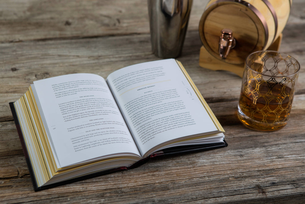 Open book on a wooden table with a glass of whiskey and a barrel in the background