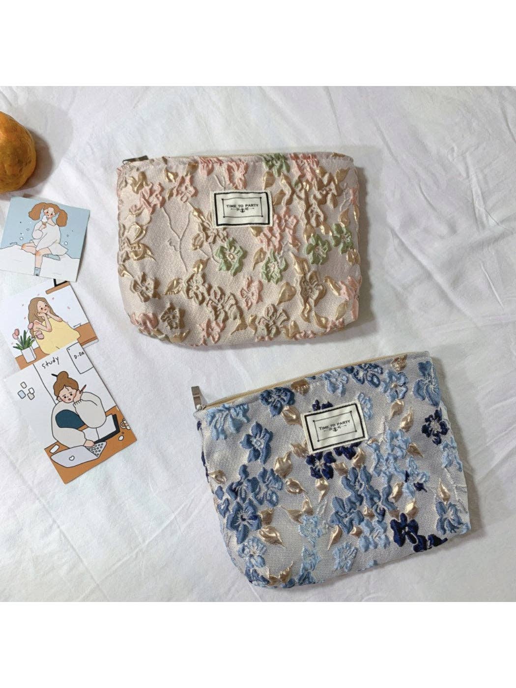 Two floral embroidered makeup pouches displayed on a white fabric background, with one in soft pink and green tones and the other in blue and gold tones. A small illustrated card is partially visible on the left.