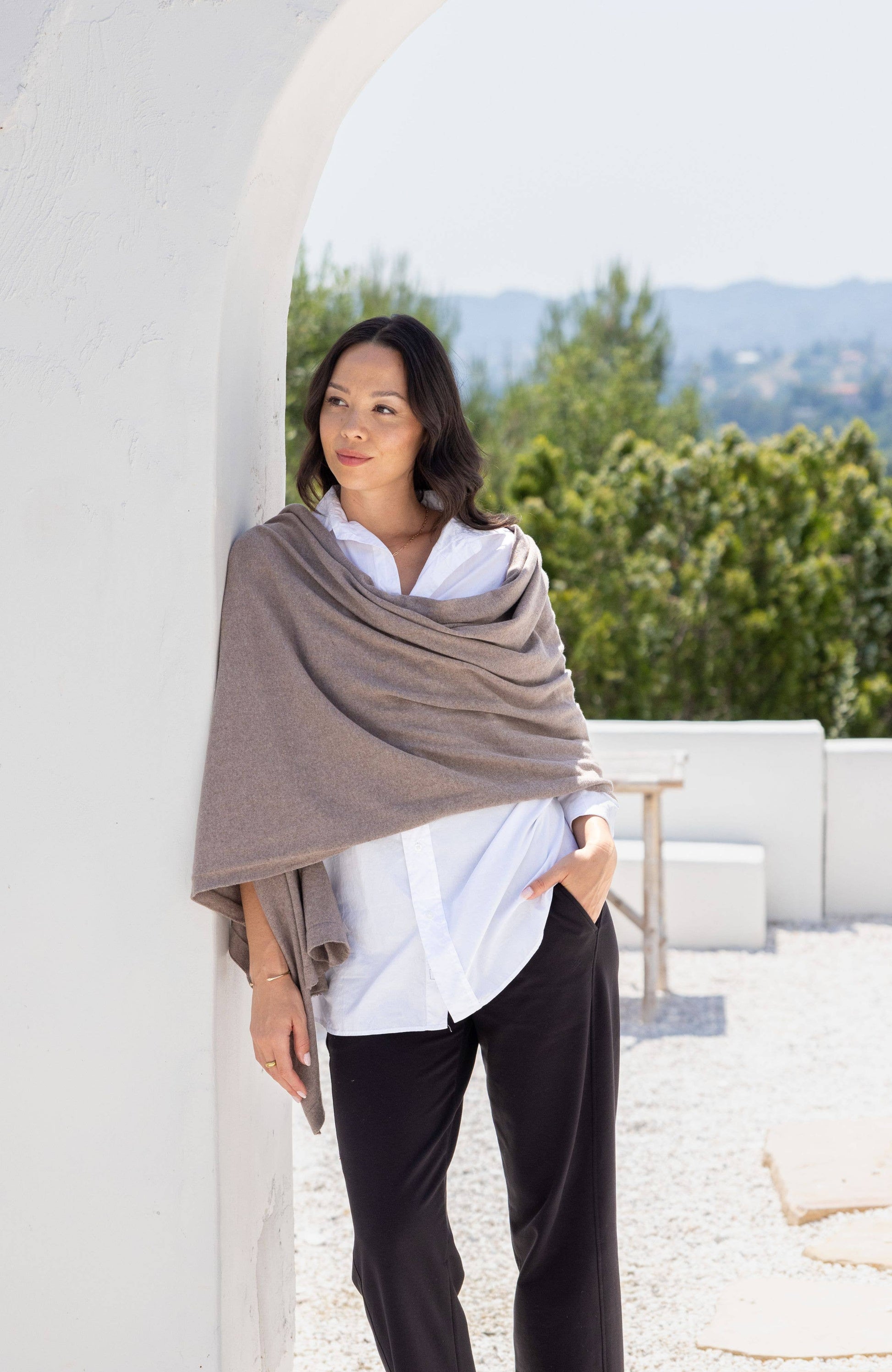 Woman wearing a beige shawl over a white shirt and black pants, standing outdoors with greenery in the background.