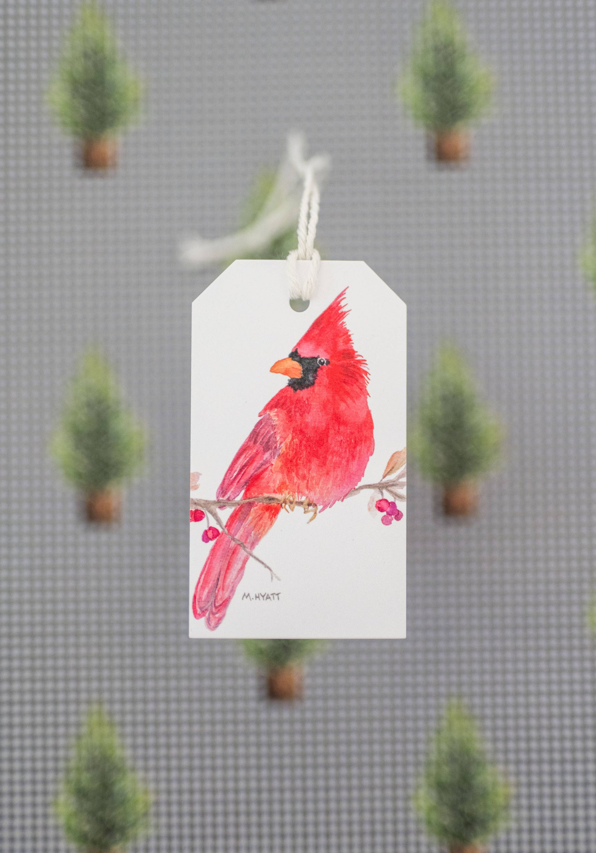 A white gift tag with a watercolor illustration of a bright red cardinal perched on a branch, hanging against a gray patterned background with blurred green pine trees.