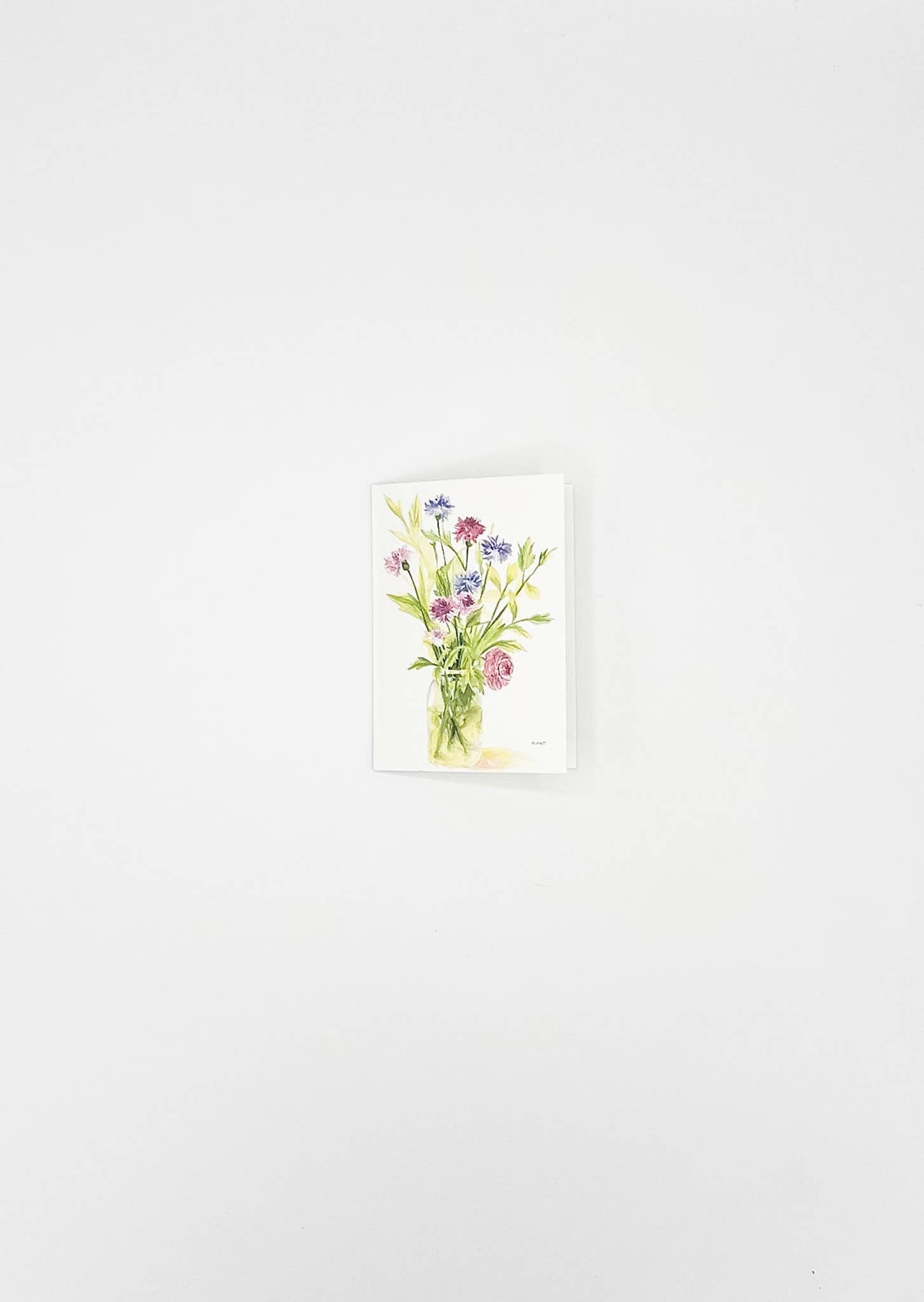 A small vertical watercolor painting of a wildflower bouquet in a clear glass vase, displayed against a plain white wall.