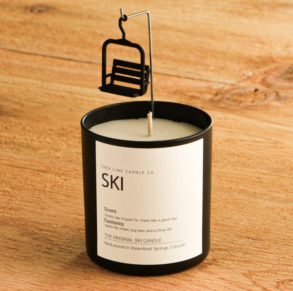 A black glass jar candle with a white label reading 'SKI' and 'FREE LINE CANDLE CO' sits on a wooden surface. A small black ski chair hangs from the candle wick by a wire loop, creating the illusion of a suspended ski lift.