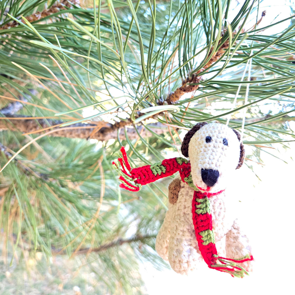 Crocheted dog ornament with a red scarf hanging on a pine branch.