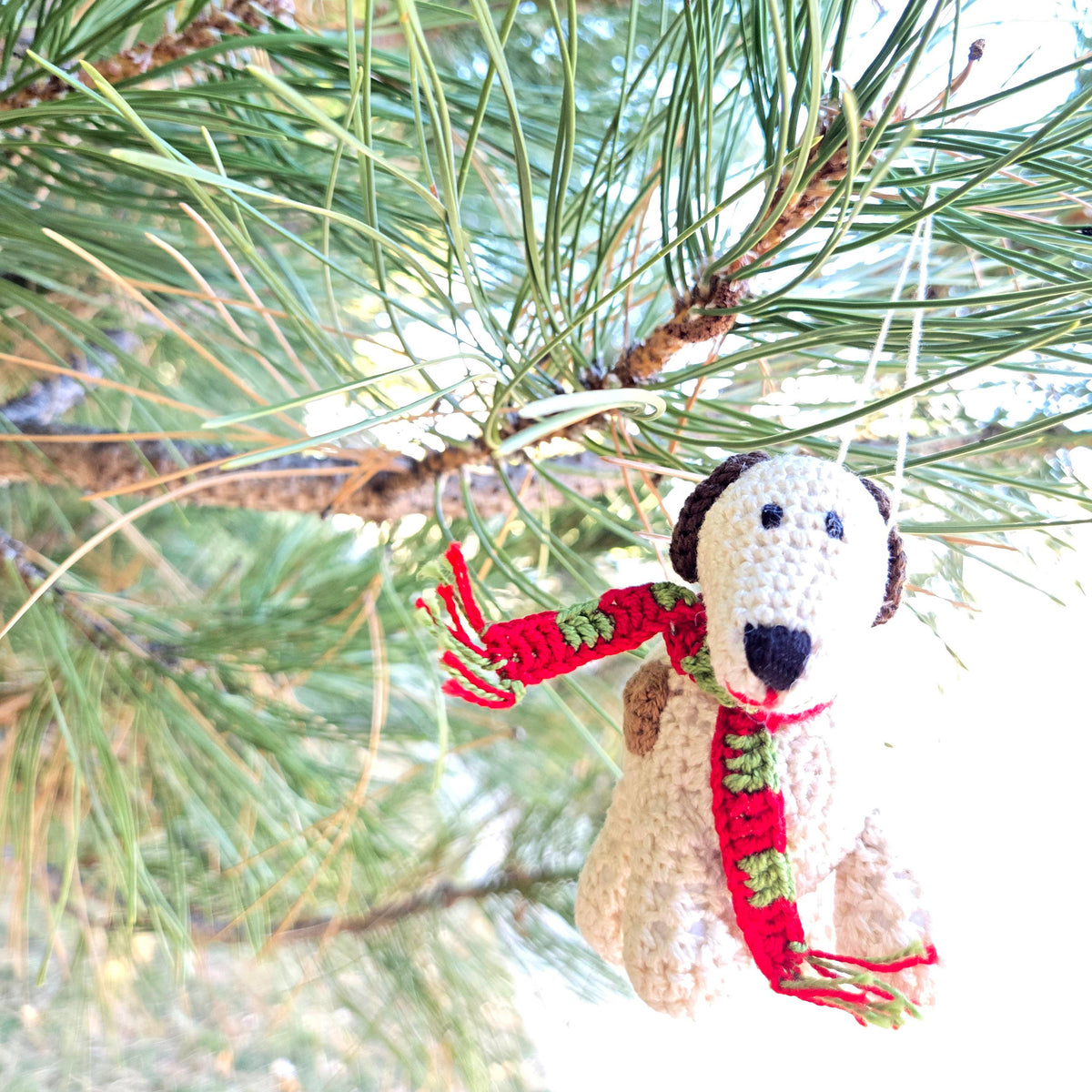 Crocheted dog ornament with a red scarf hanging on a pine branch.