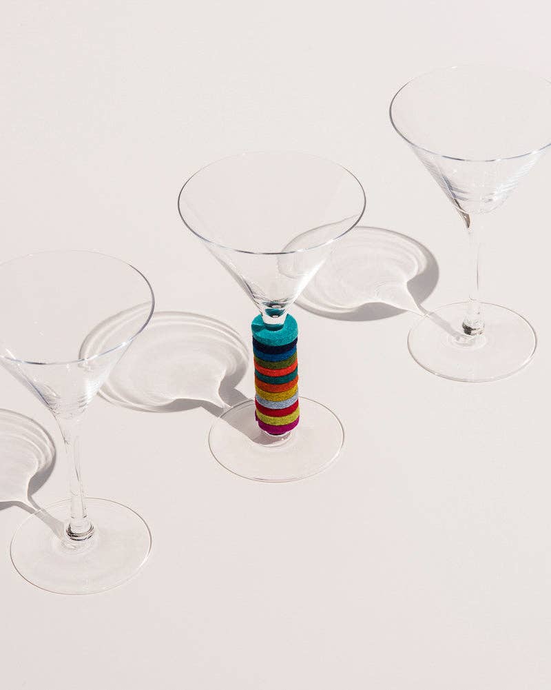 A clear stemmed cocktail glass with a colorful striped glass insert under the bowl, photographed on a clean off-white background with soft shadows.