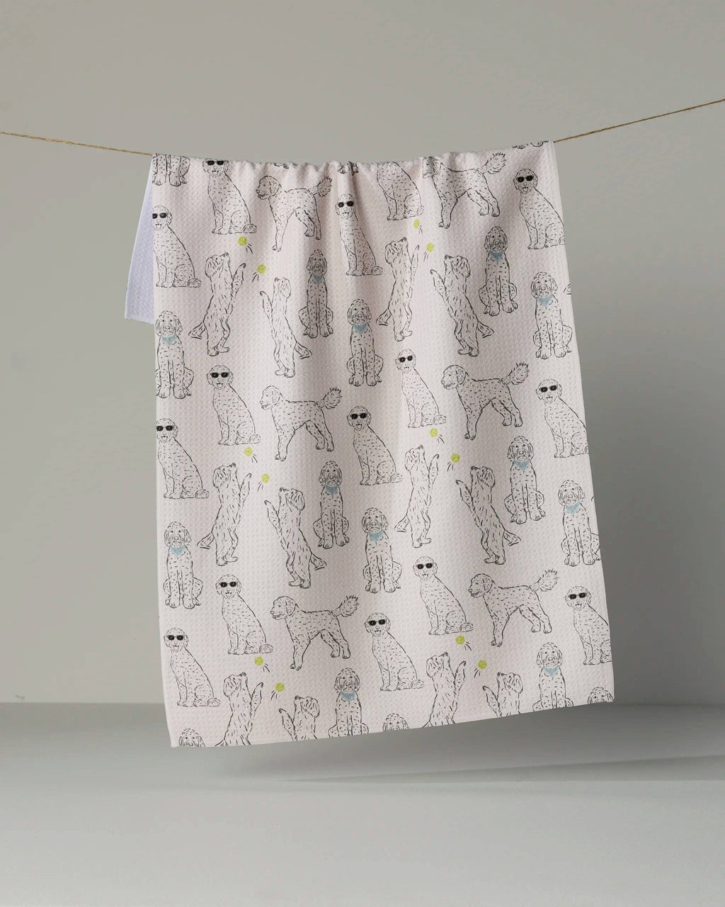 A white kitchen towel with a repeating pattern of black line drawings of dogs wearing sunglasses, hanging from a thin string against a neutral gray background.