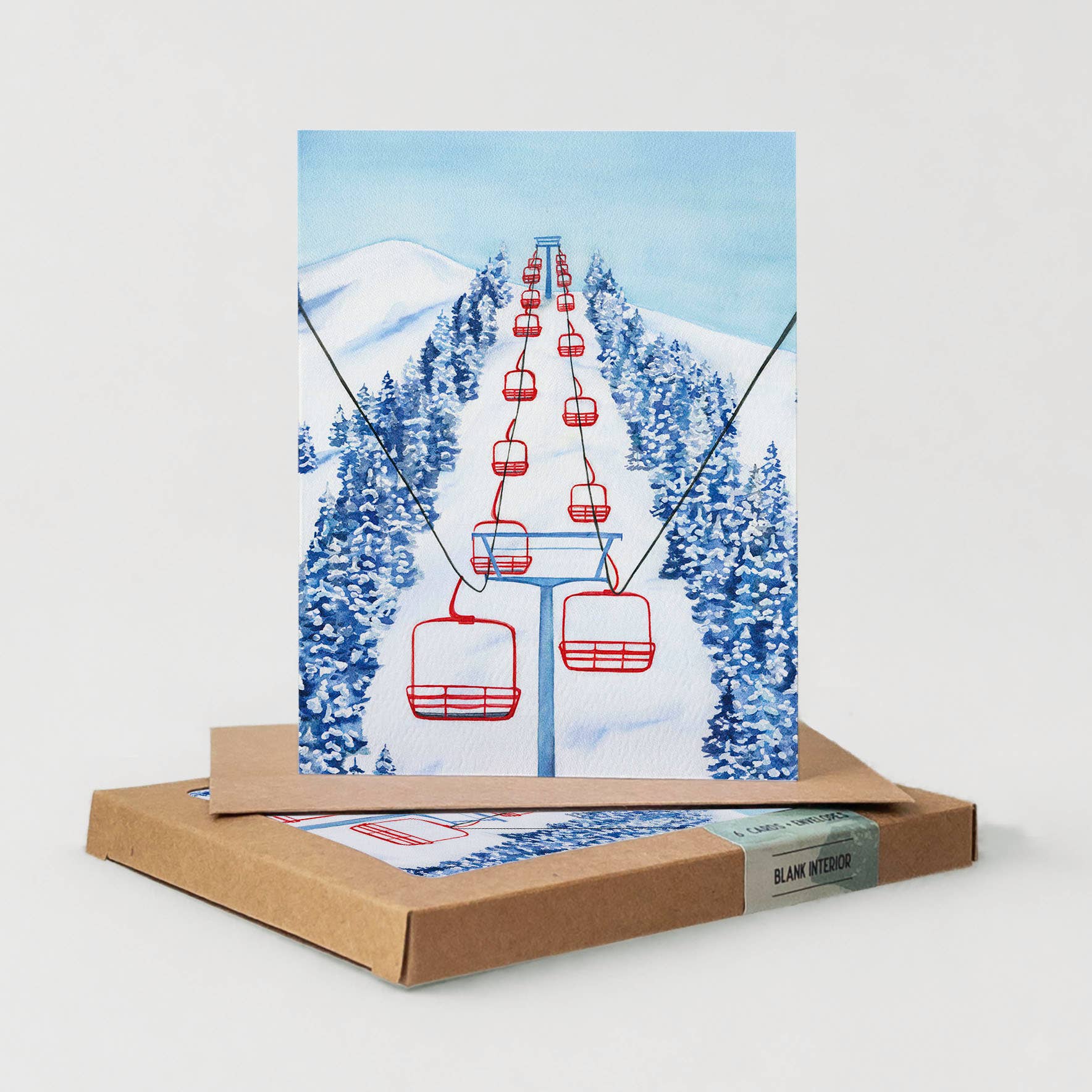 A square art print or card featuring a ski lift with red chairs over snowy mountains and blue evergreen trees, displayed upright on top of a brown kraft box. A matching printed sheet is partially visible in the box.