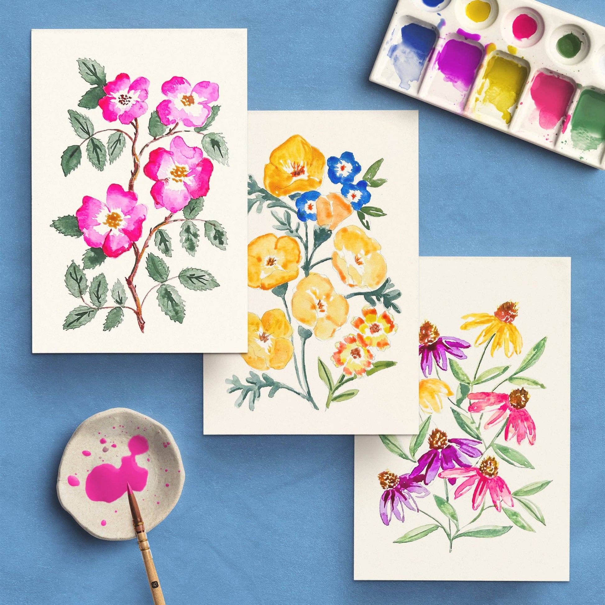 Colorful floral paintings on paper with a paint palette and brush on a blue background
