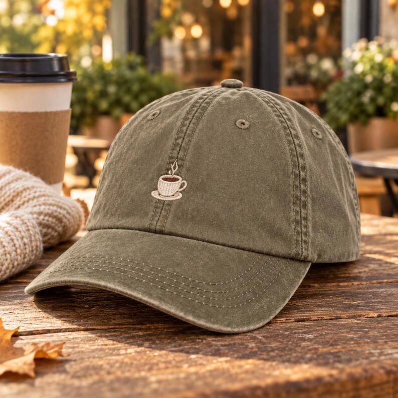 An olive green washed cotton baseball cap with a small embroidered coffee cup on the front, resting on a rustic wooden table next to a paper coffee cup and a knitted scarf in an outdoor cafe setting.