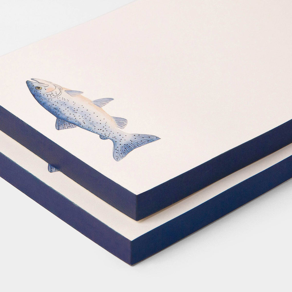 A stack of square art prints or cards with a light cream background, featuring a detailed illustration of a speckled blue-gray trout. The edges of the prints are dark navy blue, and they are arranged diagonally on a clean white background.