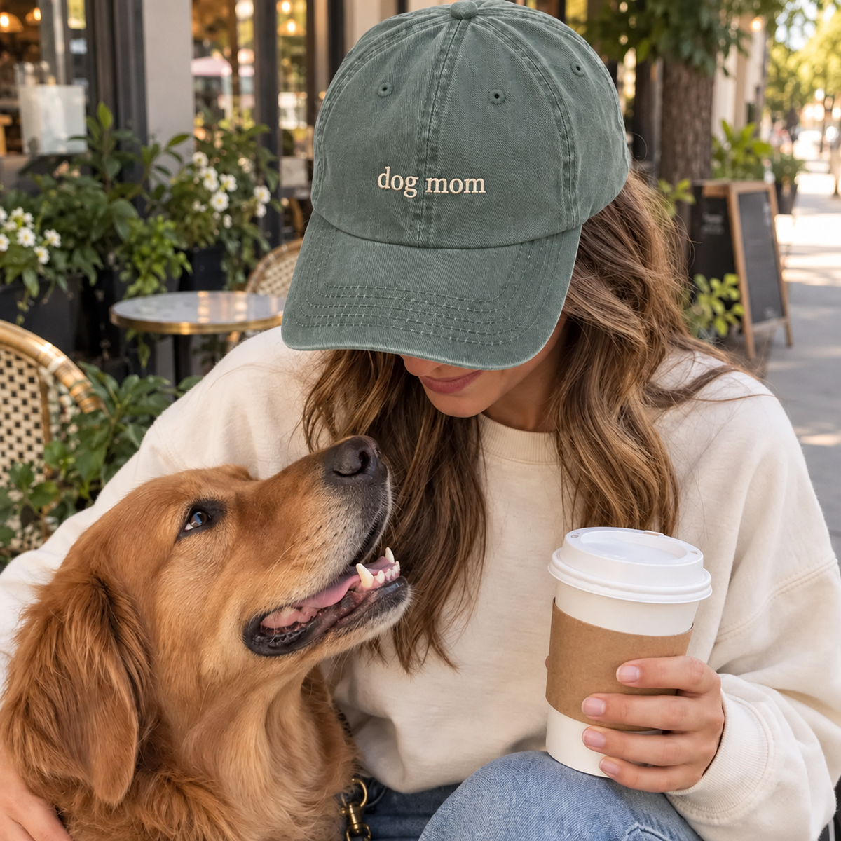 A woman wearing a muted sage green baseball cap with 'dog mom' embroidered in gold on the front, sitting outdoors with a golden retriever dog and holding a coffee cup.