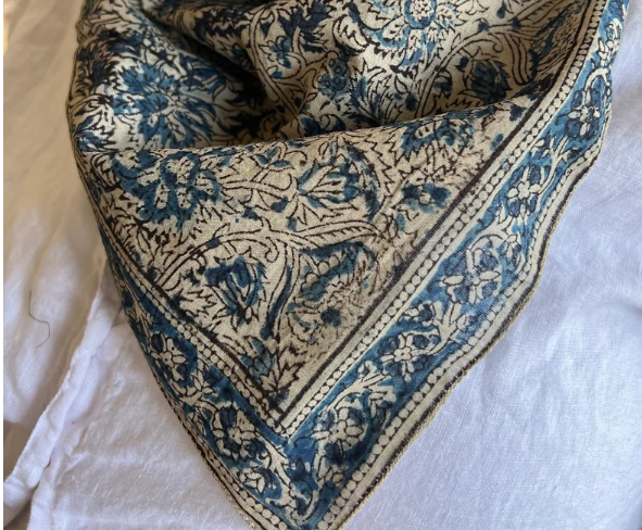 A folded square scarf or bandana featuring a detailed blue and beige floral pattern with a decorative border, displayed on a white fabric background.