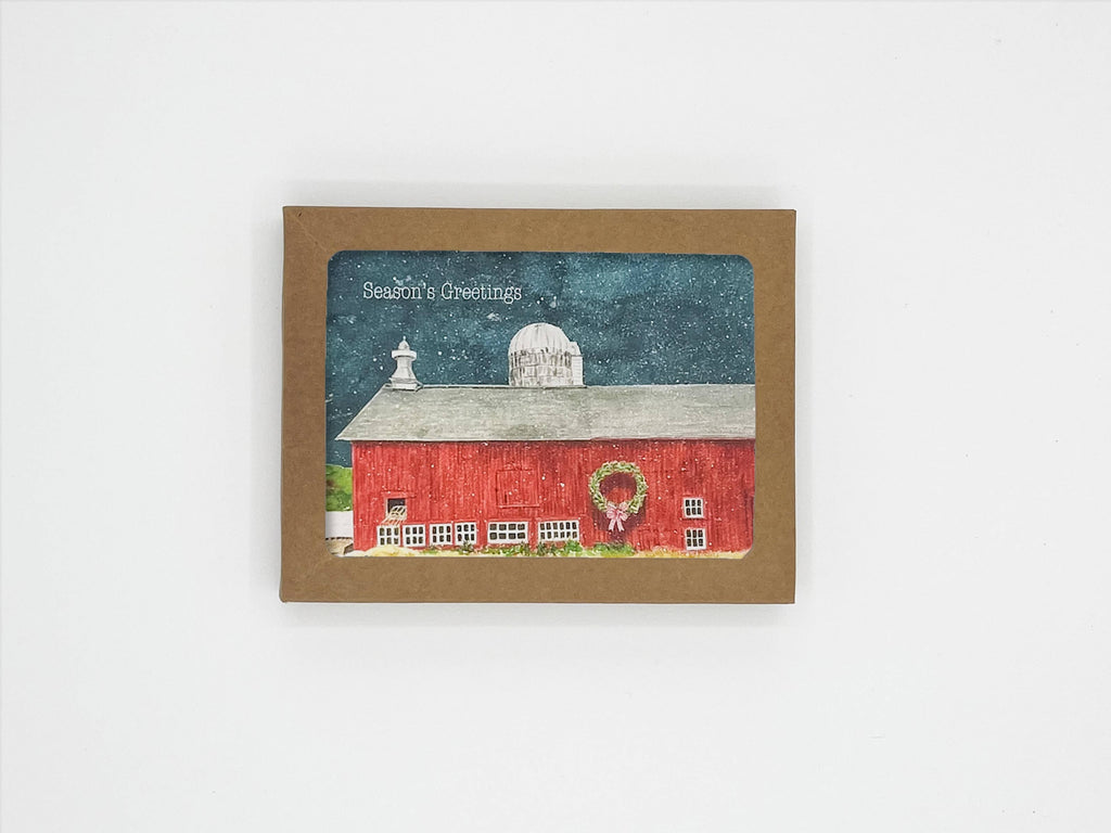A square greeting card with a brown border featuring a winter illustration of a red barn under a dark blue night sky. The card is displayed against a clean white background.