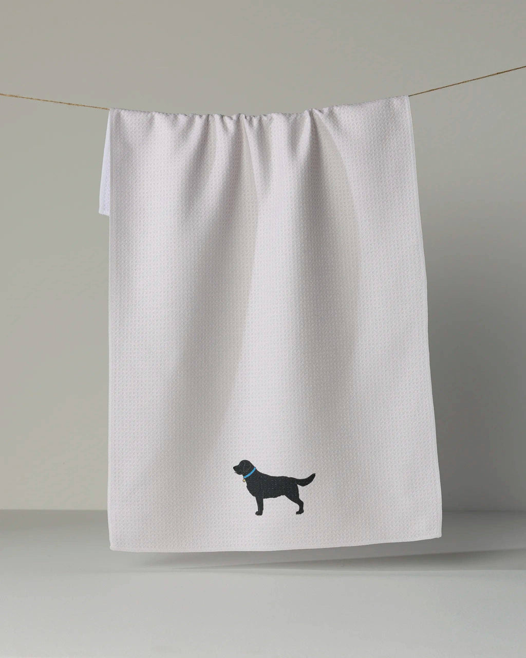 A white waffle-weave kitchen towel hanging from a string against a neutral gray background, featuring a black silhouette of a dog wearing a blue collar near the bottom.
