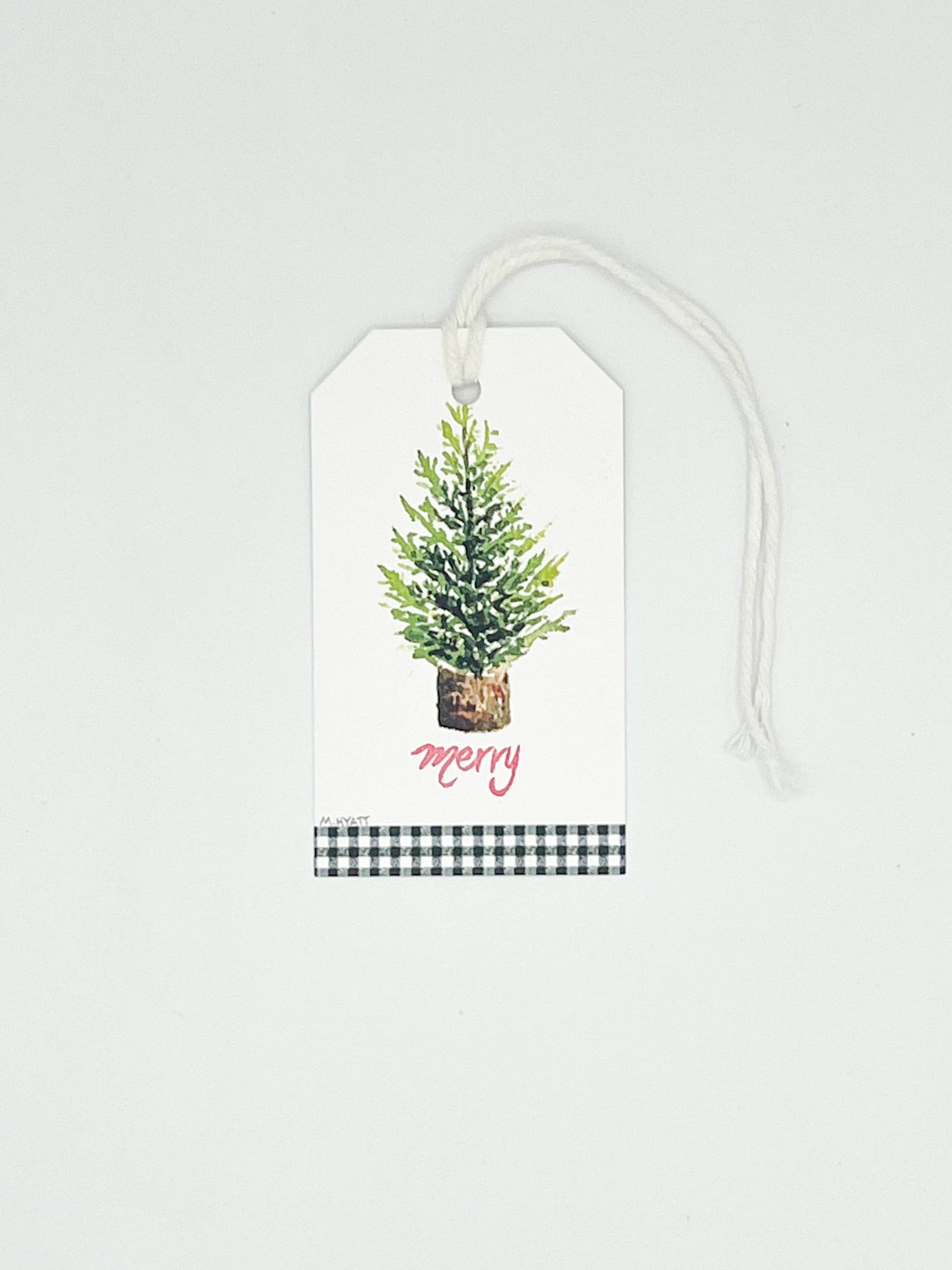 A white Christmas gift tag with a watercolor green tree in a pot, the word 'Merry' in pink script, and a black-and-white gingham ribbon at the bottom, shown on a light gray background.
