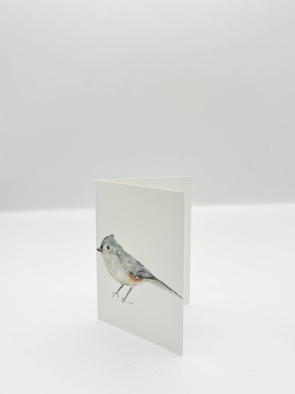 A minimalist white greeting card featuring a watercolor illustration of a small gray bird with a blue-gray crest, standing upright on a white surface against a light gray background.