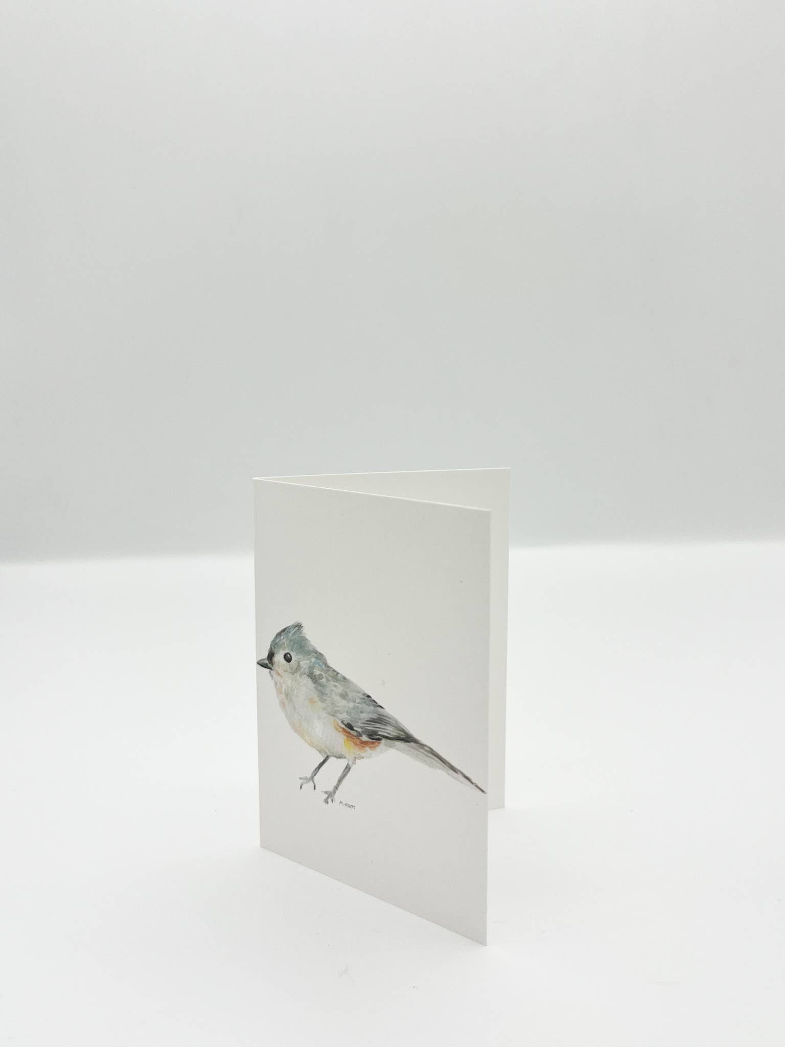 A minimalist white greeting card featuring a watercolor illustration of a small gray bird with a blue-gray crest, standing upright on a white surface against a light gray background.