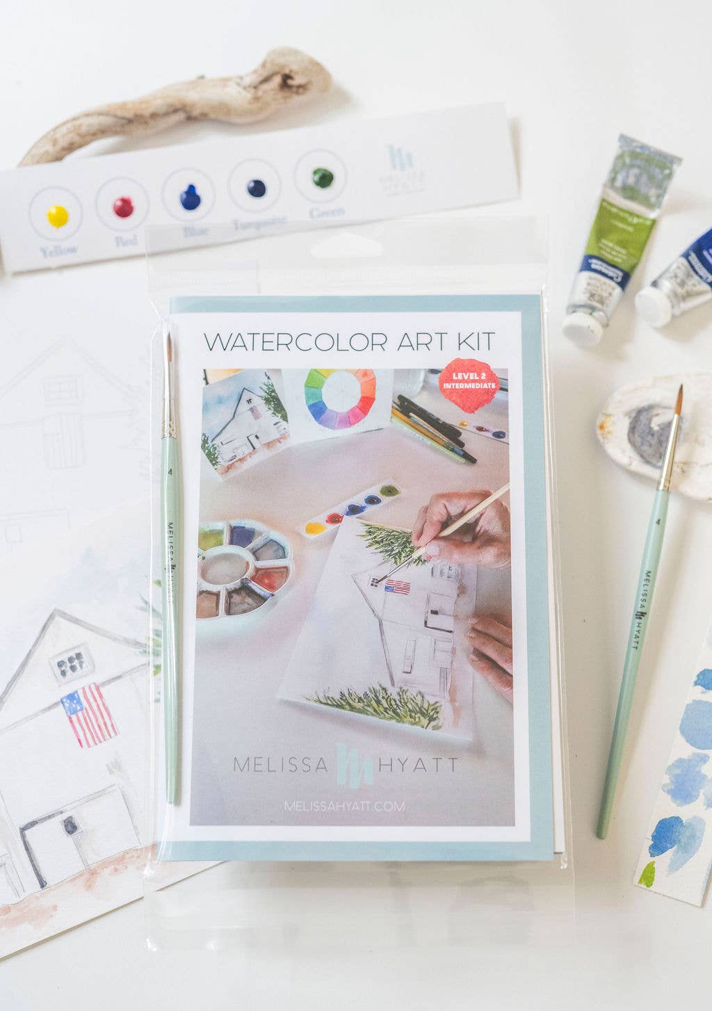 Melissa Hyatt Watercolor Art Kit in a clear plastic sleeve, displayed on a white surface with art supplies including paint tubes, a paint palette, a pencil, and a piece of driftwood.