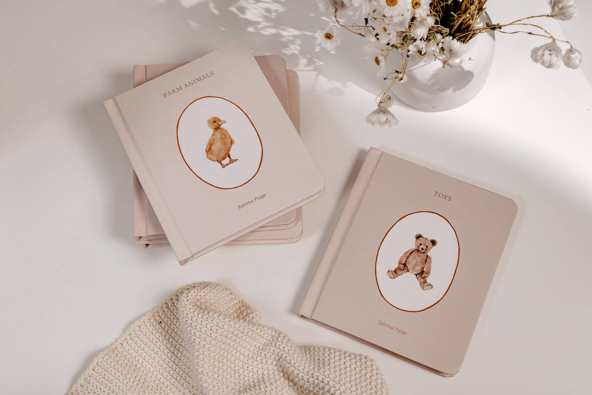 Two beige books with bear illustrations on a light surface with a vase of flowers.