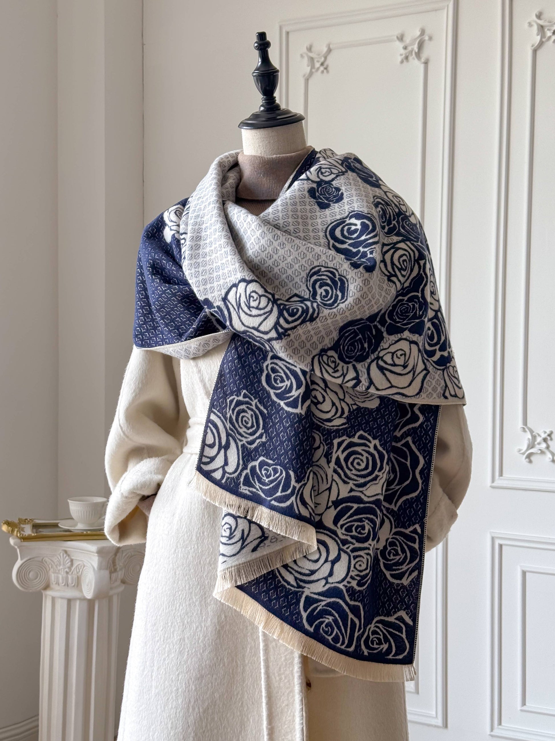 A navy blue and cream scarf with a rose pattern and fringed edges, draped on a mannequin wearing a cream coat in an elegant indoor setting.