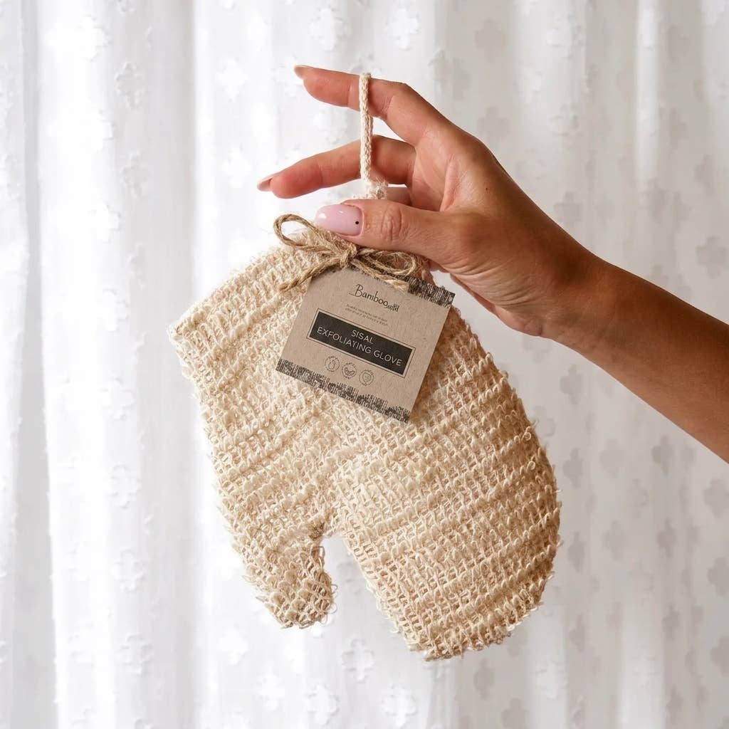 A hand holds a woven beige baby mitt with a hanging paper tag against a soft white curtain background.