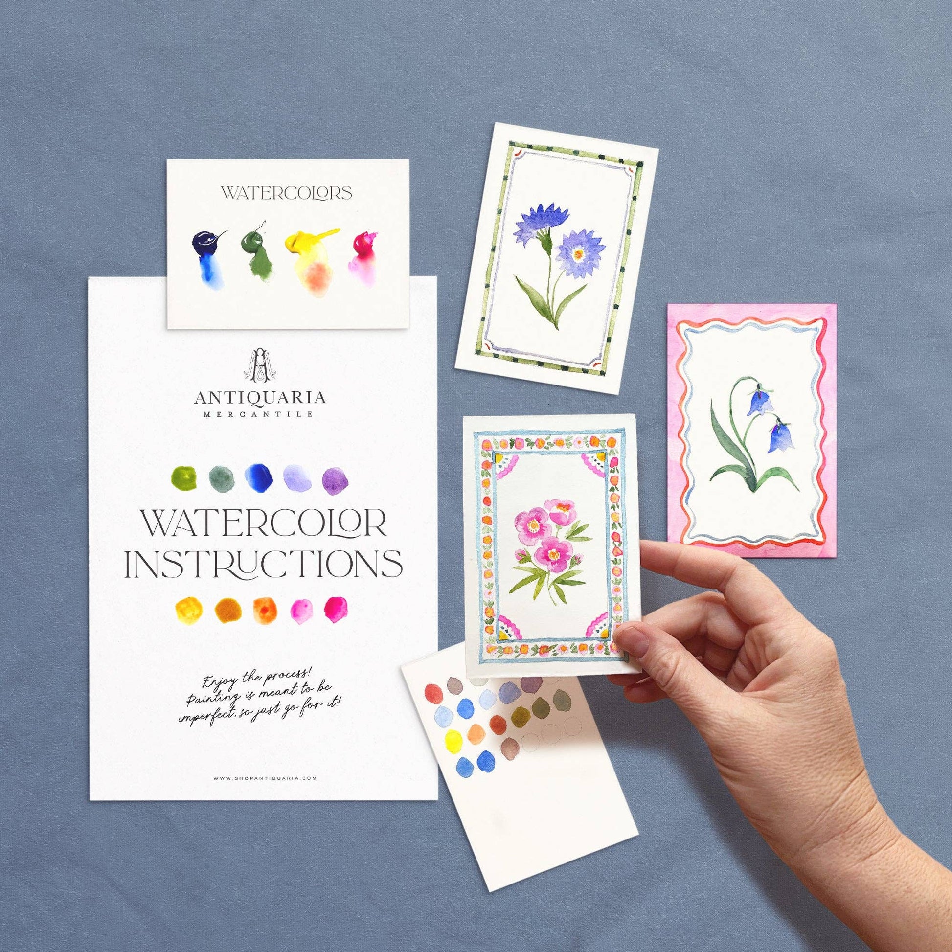 Hand holding a watercolor card with floral design, surrounded by other cards and an instruction manual on a blue background.