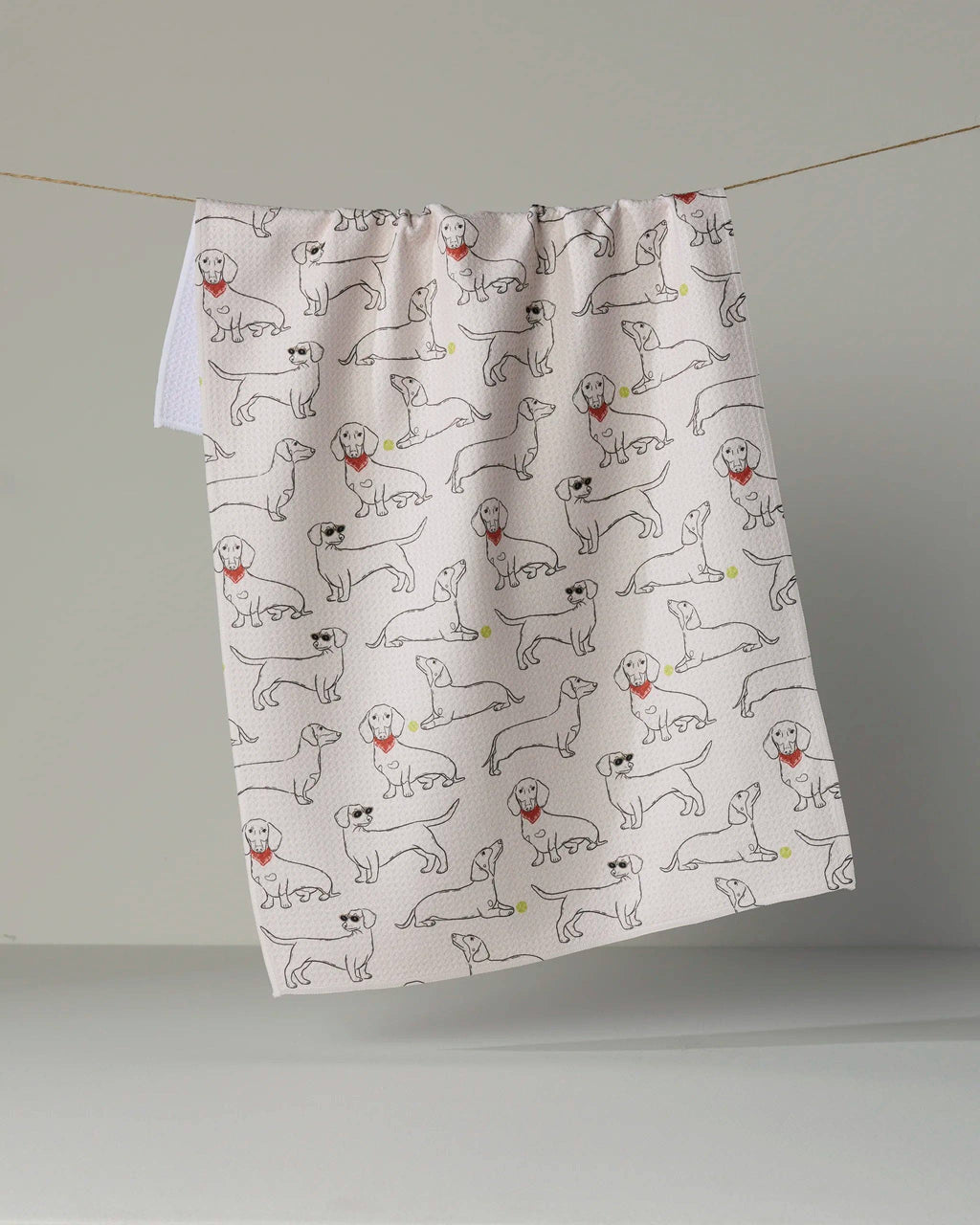 A white kitchen tea towel with a repeating pattern of hand-drawn black line drawings of dogs, some wearing red neckties, hanging from a string against a neutral gray background.