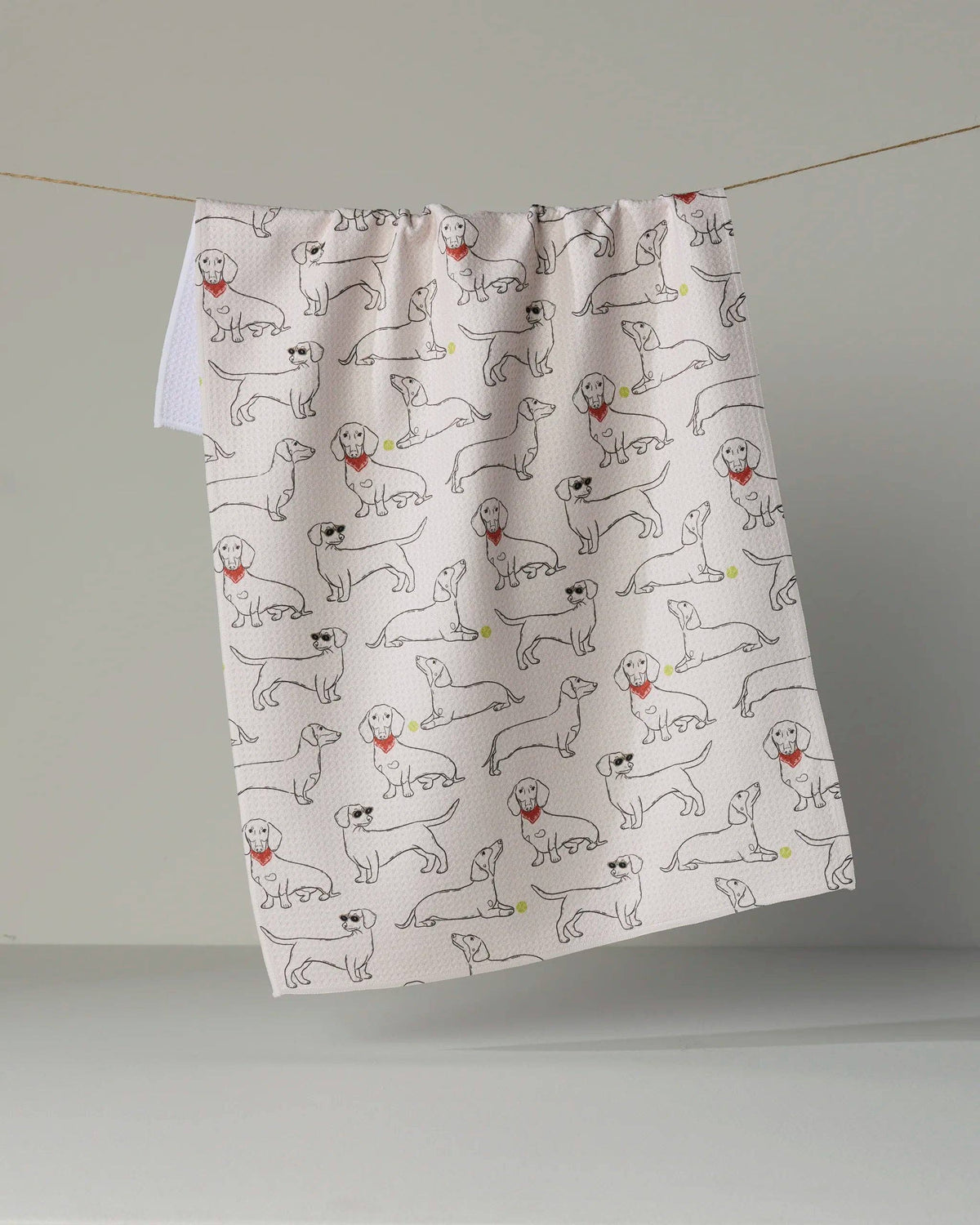 A white kitchen tea towel with a repeating pattern of hand-drawn black line drawings of dogs, some wearing red neckties, hanging from a string against a neutral gray background.