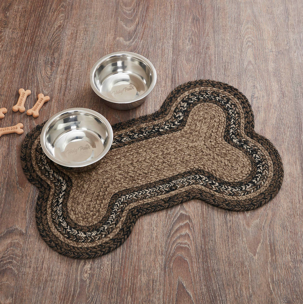A bone-shaped braided pet mat in brown and beige tones, shown on a wood floor with two stainless steel dog bowls and a few bone-shaped treats nearby.