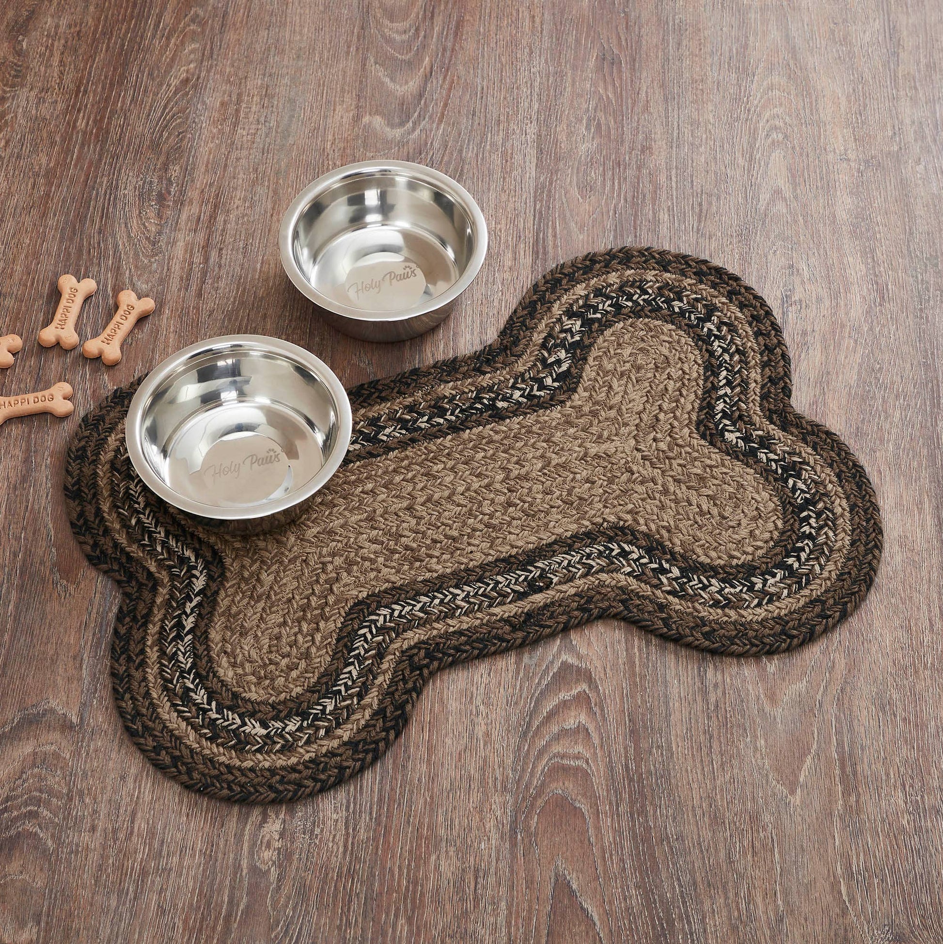 A bone-shaped braided pet mat in brown and beige tones, shown on a wood floor with two stainless steel dog bowls and a few bone-shaped treats nearby.