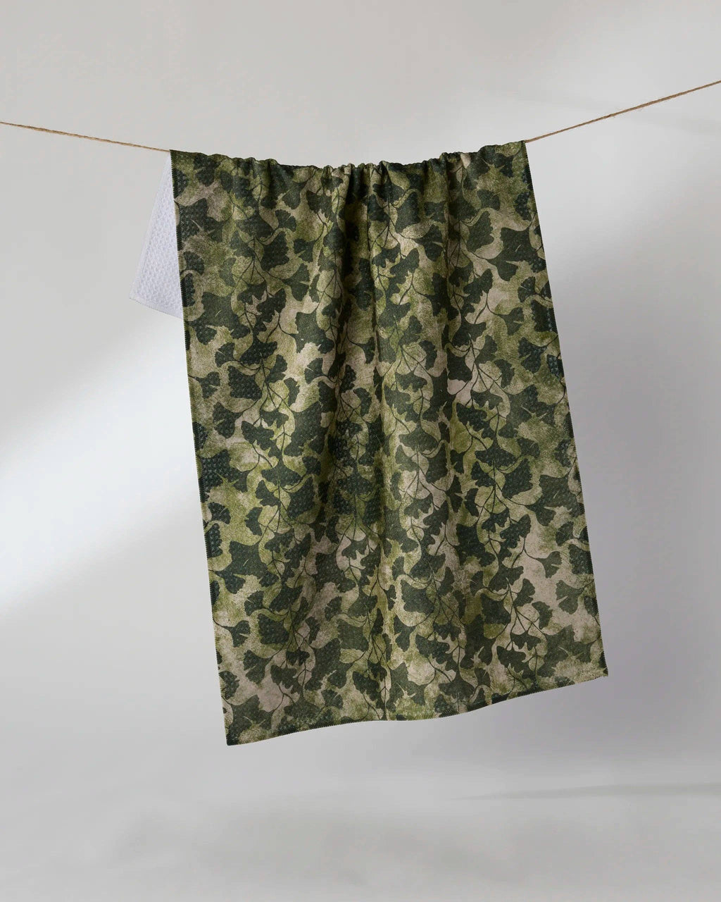 A single curtain panel with an allover ivy camouflage pattern, hanging from a thin wire on a plain light gray studio background.