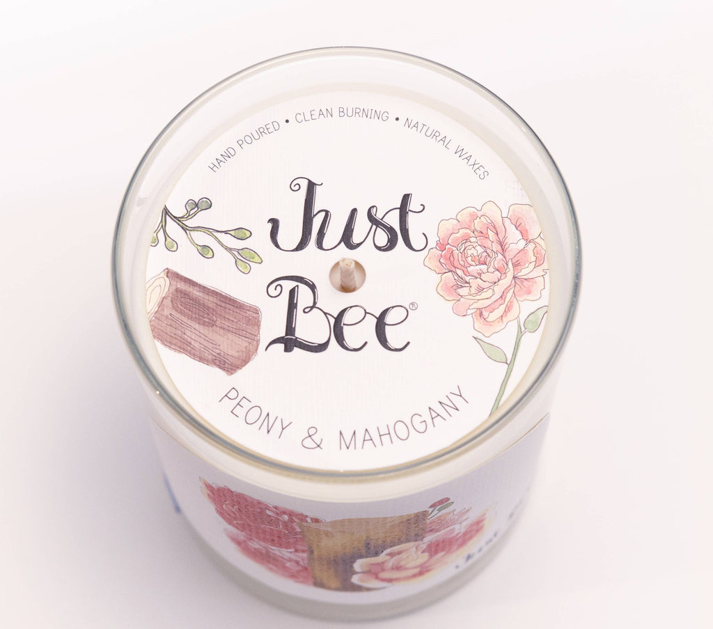 A glass jar candle viewed from above, showing the white lid label with the brand name 'Just Bee' and the scent name 'Peony & Mahogany' in black and gray text. The label features illustrations of a pink peony flower, a pink-and-brown wax melt cube, and green leaves on a white background.