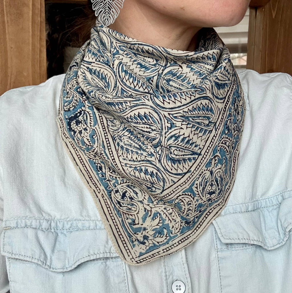 A close-up of a person wearing a square bandana-style neck gaiter with an intricate paisley and floral block-print pattern in blue and beige, styled over a light blue denim shirt.