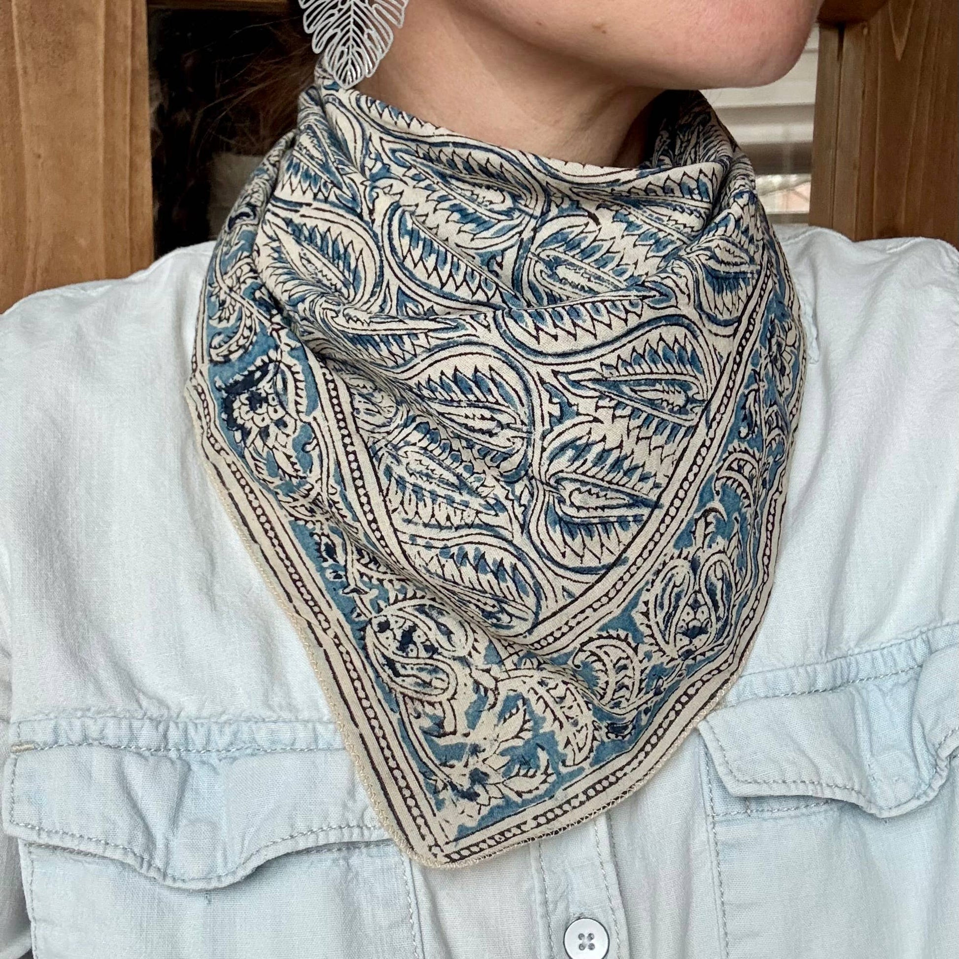 A close-up of a person wearing a square bandana-style neck gaiter with an intricate paisley and floral block-print pattern in blue and beige, styled over a light blue denim shirt.