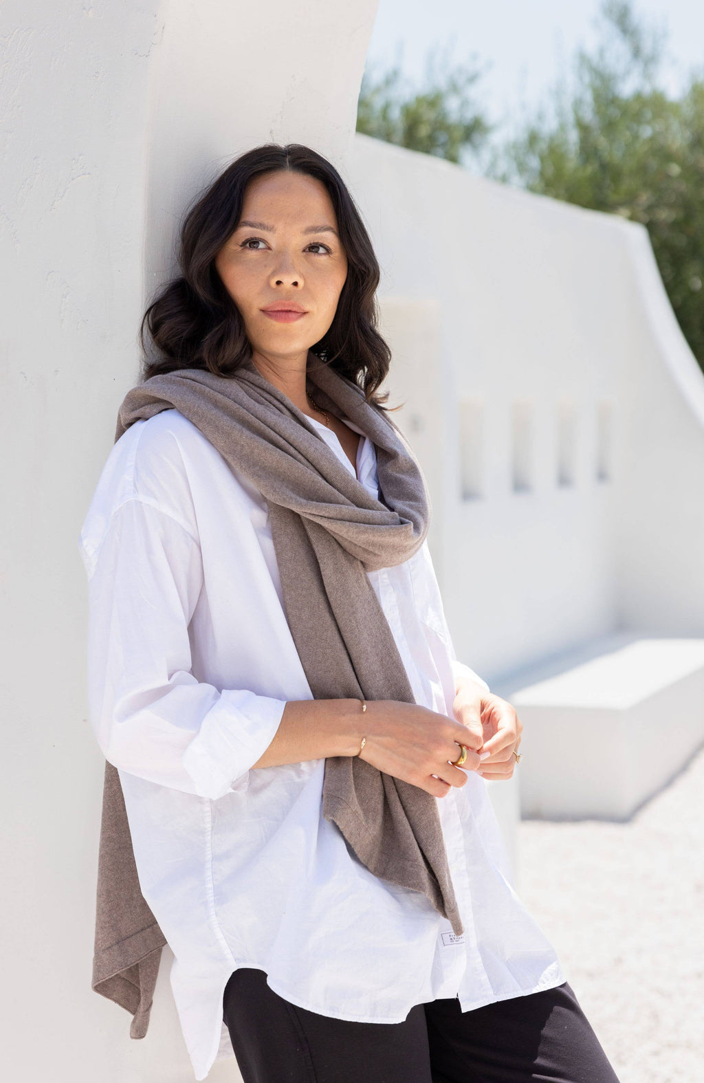Woman wearing a white shirt and brown scarf against a white wall with greenery.