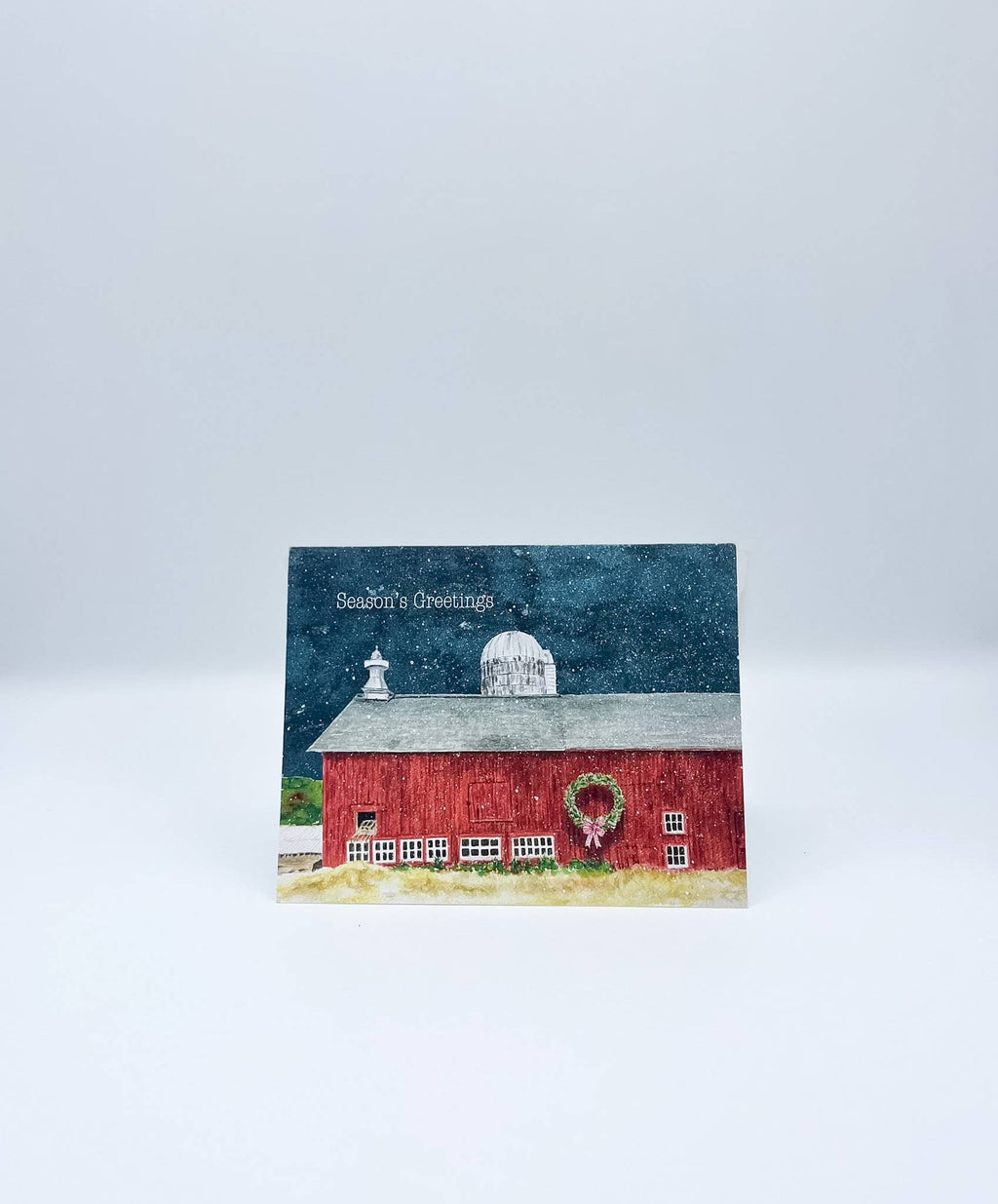 A square Christmas greeting card featuring a painted red barn with a white dome, a green wreath, and the text 'Season's Greetings' on a snowy night sky, displayed on a light gray background.