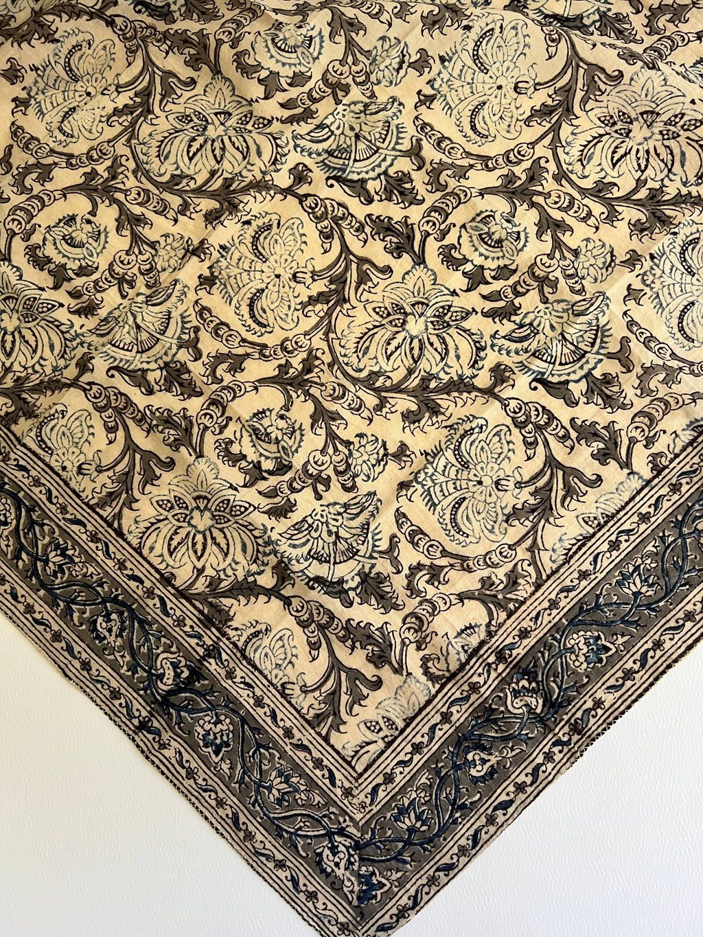 A close-up of an ornate patterned textile or rug corner, showing a cream background with dark brown and muted blue-gray floral and scroll motifs and a decorative border.