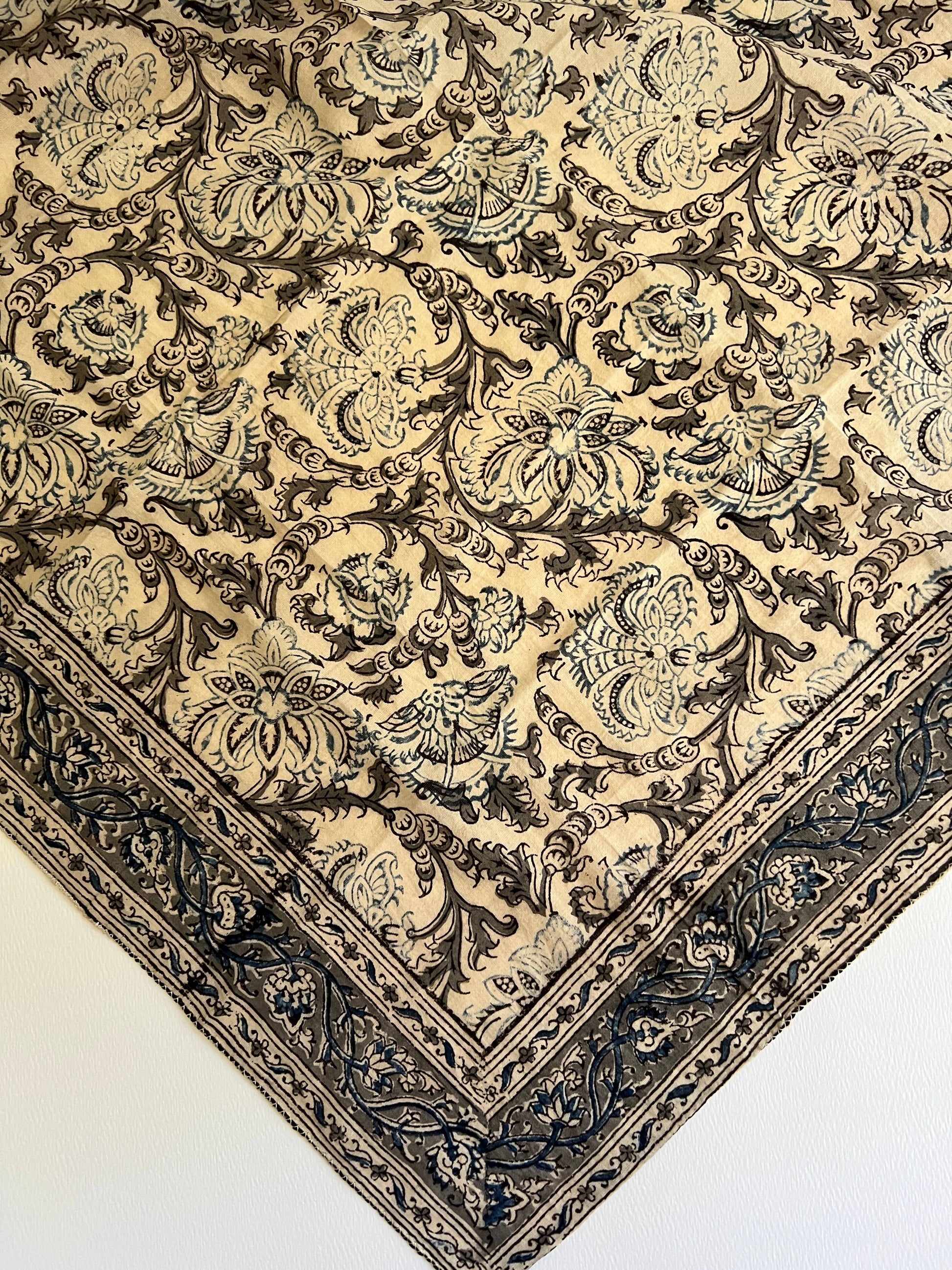 A close-up of an ornate patterned textile or rug corner, showing a cream background with dark brown and muted blue-gray floral and scroll motifs and a decorative border.