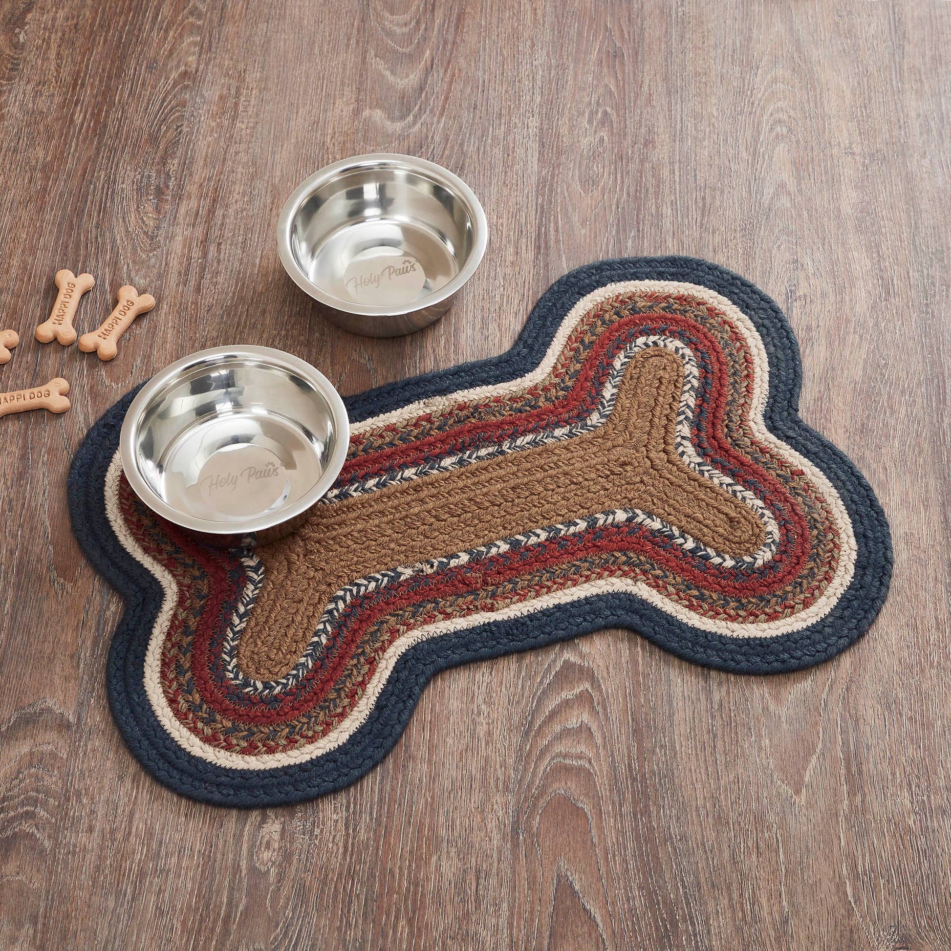 A braided bone-shaped pet mat in brown, beige, red, and navy, shown on a wood floor with two stainless steel dog bowls and small bone-shaped toys.