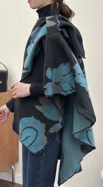 A woman wearing a black turtleneck sweater and blue jeans, draped in a large black knit poncho with a blue floral pattern, standing indoors.
