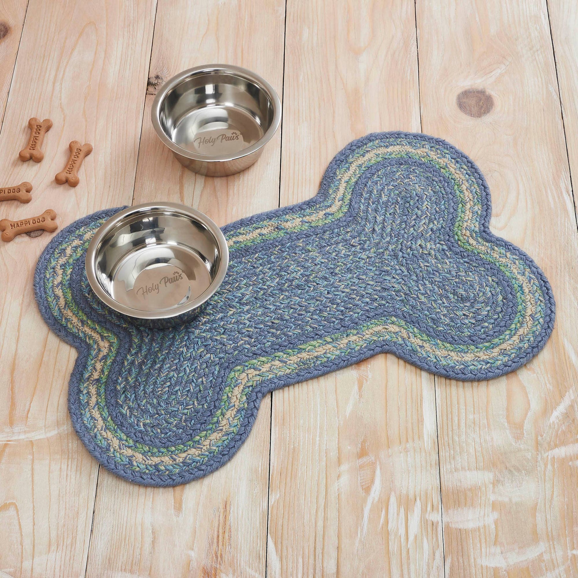 A bone-shaped braided pet mat in blue, green, and beige on a light wood plank floor, with two stainless steel bowls and a few dog treats placed nearby.