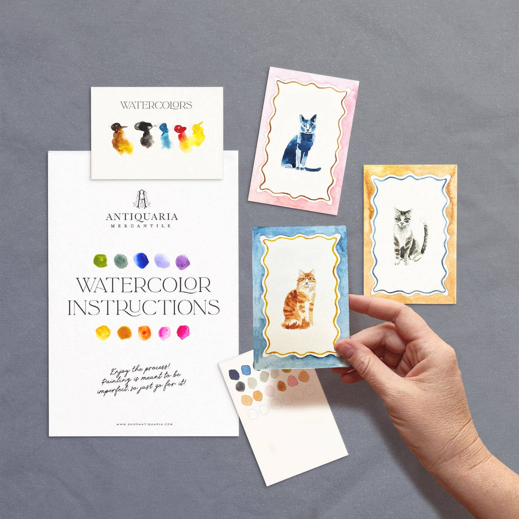 Hand holding a watercolor card with floral design, surrounded by other cards and an instruction manual on a blue background.